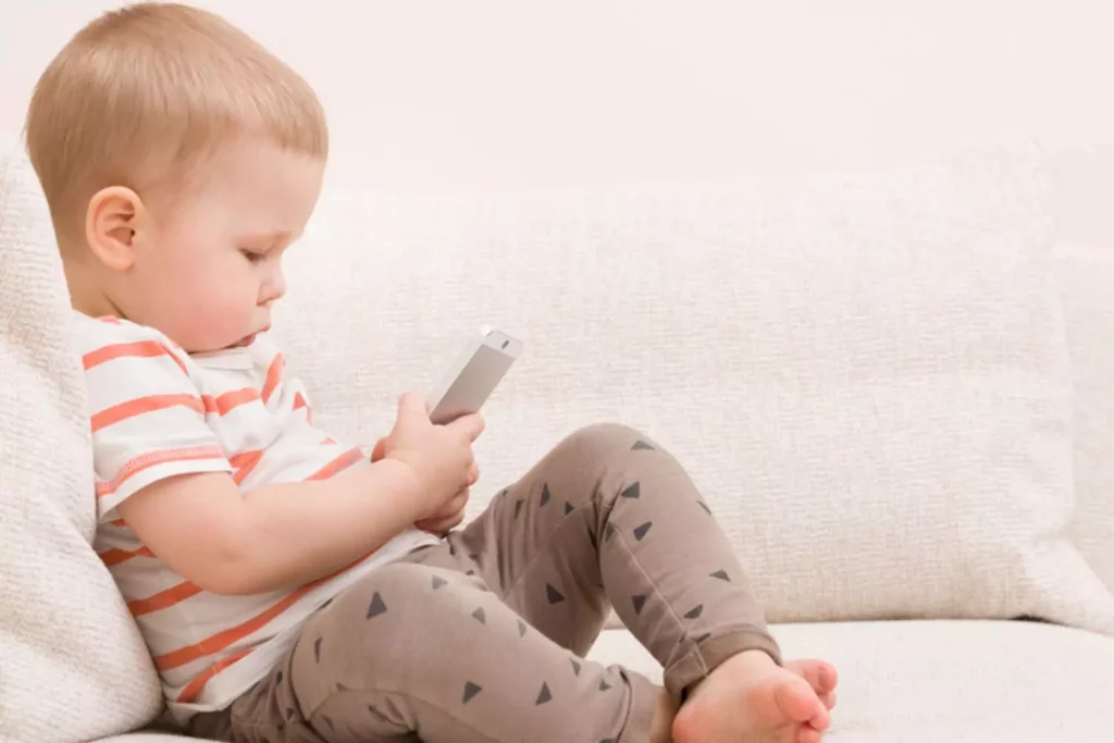 Addicted to screens- a toddler addicted to screens is sitting on the couch holding a smartphone