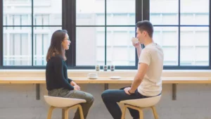 Photo of a woman initiating a conversation with her husband as they sit at a table in front of large windows while the man sips coffee and listens to her.
