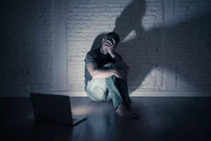 A man hides his face in shame from a computer screen in a dark room. People may use sex and pornography to activate a “pleasure system high” that can lead to a deeper progression of porn addiction.